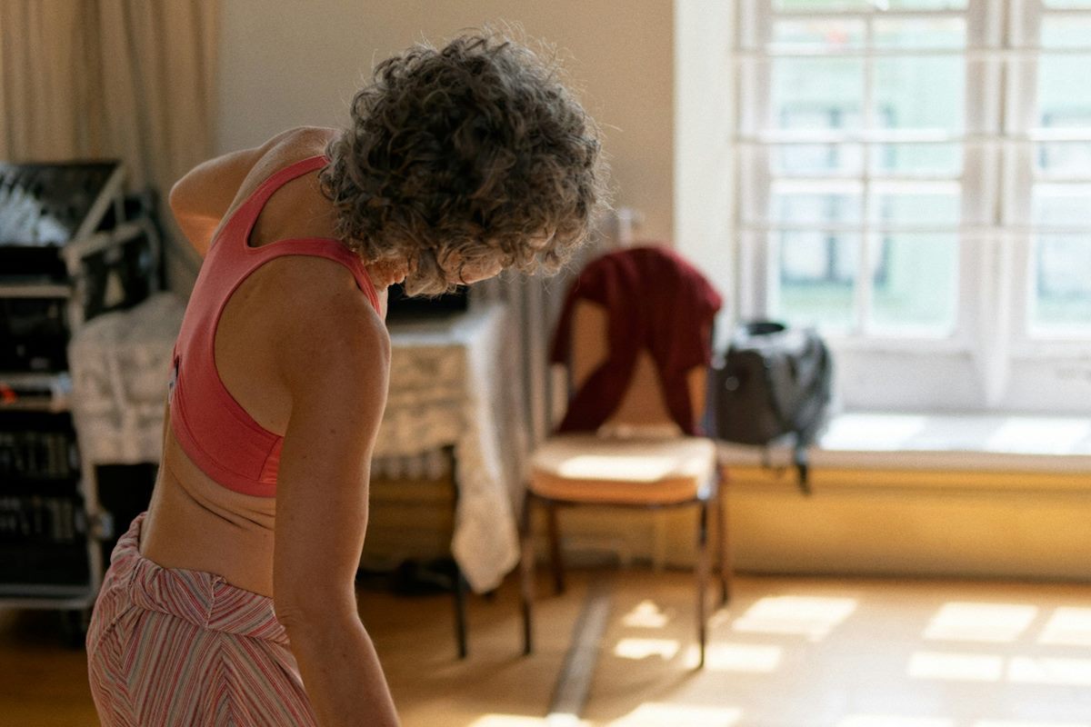 A grey-haired woman doing a gentle neck and shoulder stretch in a sunlit room