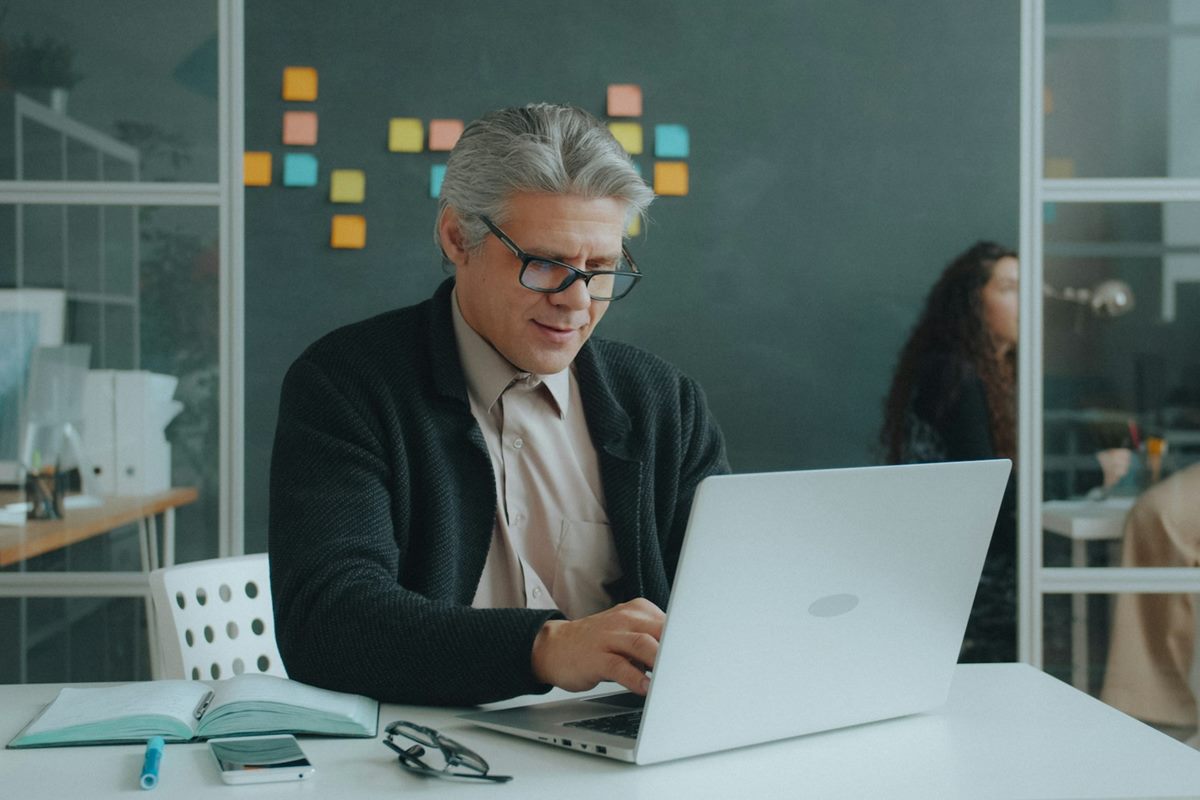 A grey-haired man in glasses working at a laptop, sitting at his desk for long hours